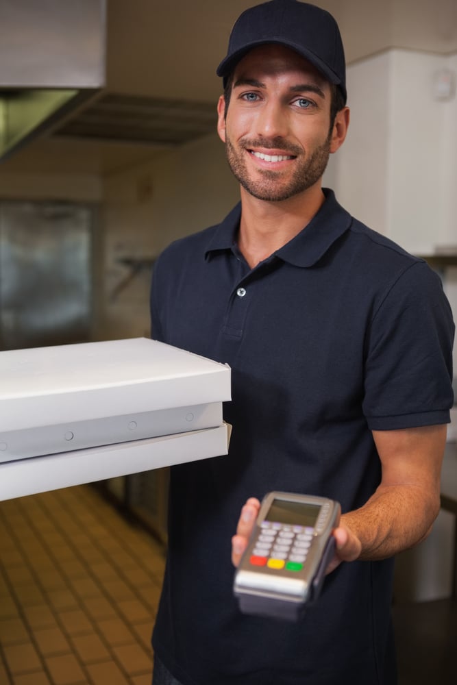 Happy pizza delivery man showing credit card machine in a commercial kitchen