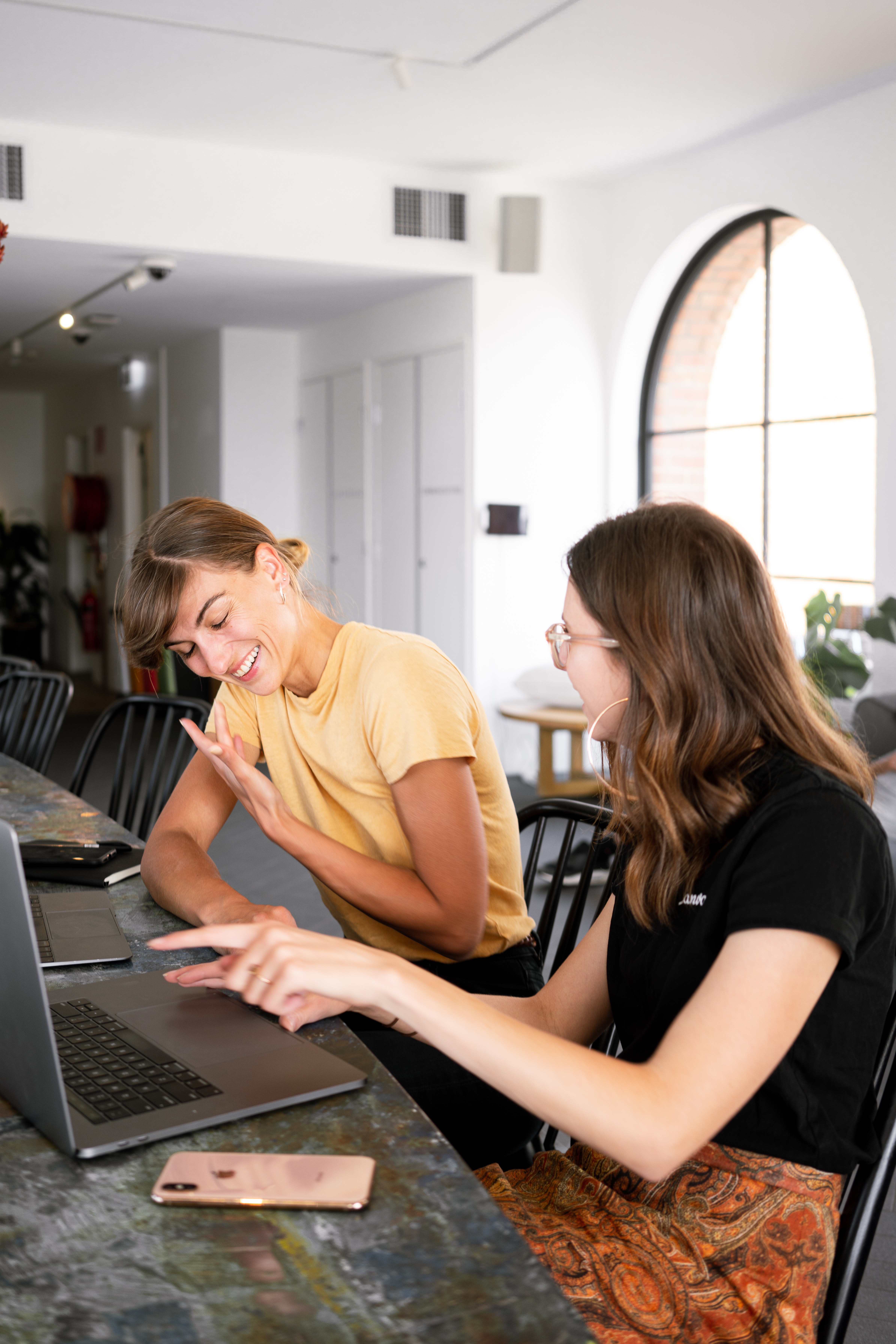 Two women sitting at a table in an office smiling at each other.